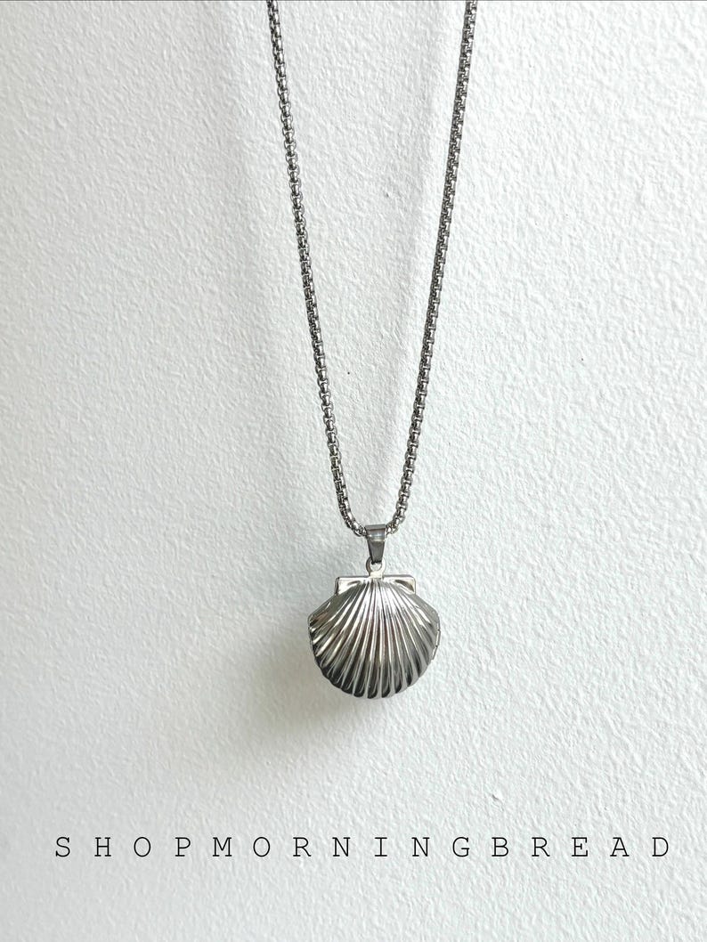 May include: A silver-colored necklace with a seashell pendant. The pendant is a detailed, ridged seashell design. The necklace hangs against a white textured background. The text "SHOP MORNING BREAD" is at the bottom of the image.