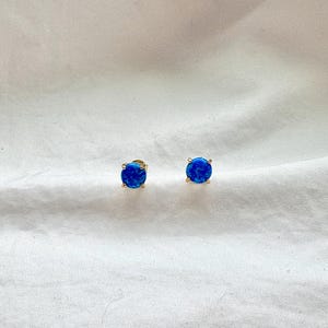 May include: A pair of stud earrings featuring round, vibrant blue gemstones set in gold-colored metal. The earrings are displayed on a white, textured surface, showcasing their color and design. These earrings are a classic jewelry accessory.