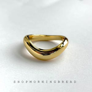May include: A gold ring with a smooth, curved design. The ring has a polished finish and is set against a white background. The text "SHOP MORNING BREAD" is printed below the ring.