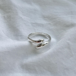Könnte beinhalten: Ein silberner Ring mit einem einzigartigen Design. Der Ring hat ein glattes, poliertes Band, das sich oben krümmt und überlappt und eine Tropfenform bildet. Der Ring ist vor einem weißen Hintergrund platziert.