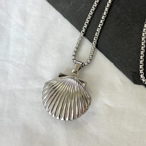 May include: A silver-colored seashell pendant necklace. The pendant is a detailed replica of a scallop shell, attached to a chain necklace. The necklace is displayed on a white surface.
