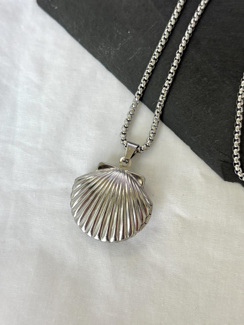 May include: A silver-colored seashell pendant necklace. The pendant is a detailed replica of a scallop shell, attached to a chain necklace. The necklace is displayed on a white surface.