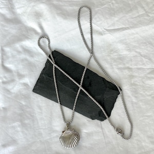 May include: A silver-colored necklace featuring a seashell pendant. The pendant has a detailed, ridged design. The necklace chain is a box chain style. The jewelry is displayed on a black, rectangular stone against a white background.
