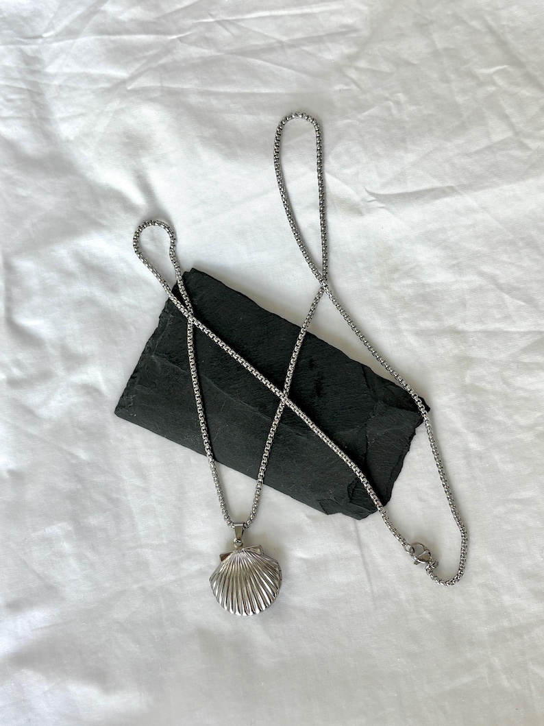 May include: A silver-colored necklace featuring a seashell pendant. The pendant has a detailed, ridged design. The necklace chain is a box chain style. The jewelry is displayed on a black, rectangular stone against a white background.