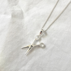 May include: A silver necklace with a pendant in the shape of scissors. The scissors pendant has open blades and finger holes, suspended from a fine chain. The necklace is shown on a white, textured background.