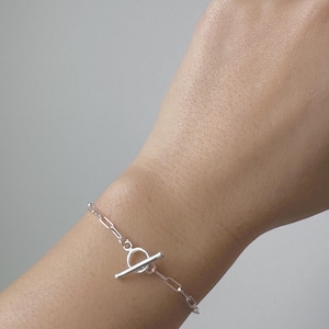 May include: A silver bracelet with a toggle clasp closure. The bracelet features a delicate chain link design. The toggle clasp is a bar that fits through a circular loop to secure the bracelet. The bracelet is worn on a wrist.