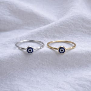 May include: Two delicate rings, one silver and one gold, each feature a small, round charm with a blue and black evil eye design. The rings are set against a white, textured background, showcasing the jewelry's simple elegance.