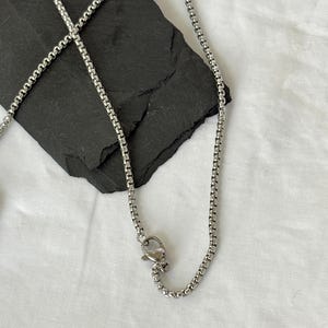 May include: A silver-colored chain necklace with a box chain design. The necklace is displayed on a black, textured surface against a white background. The chain has a lobster clasp closure.