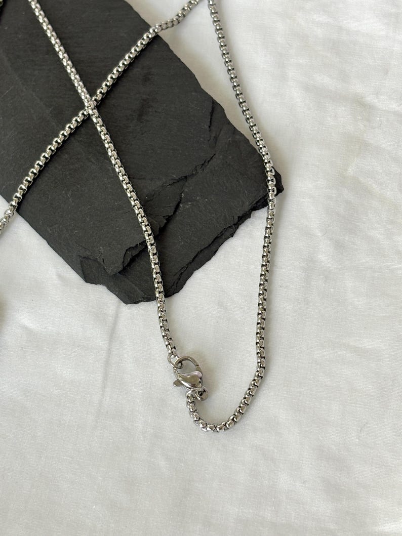 May include: A silver-colored chain necklace with a box chain design. The necklace is displayed on a black, textured surface against a white background. The chain has a lobster clasp closure.