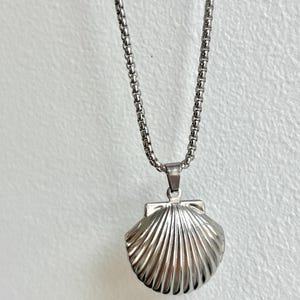 May include: A silver-colored seashell pendant necklace. The pendant is a detailed, three-dimensional seashell design with a textured surface. The necklace chain is a delicate, linked design. The background is a plain, off-white color.