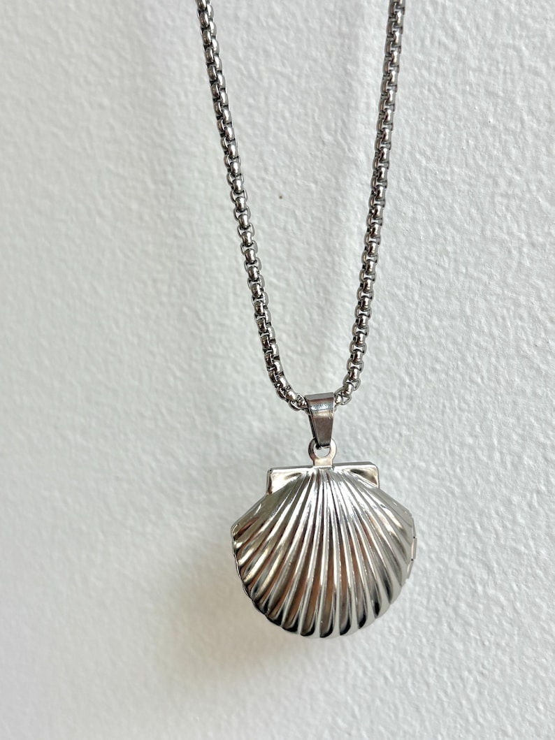 May include: A silver-colored seashell pendant necklace. The pendant is a detailed, three-dimensional seashell design with a textured surface. The necklace chain is a delicate, linked design. The background is a plain, off-white color.