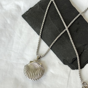 May include: A silver-colored seashell pendant necklace. The pendant is a detailed, three-dimensional scallop shell design. The necklace chain is a delicate, box-style chain. The pendant is displayed on a black, textured surface against a white background.