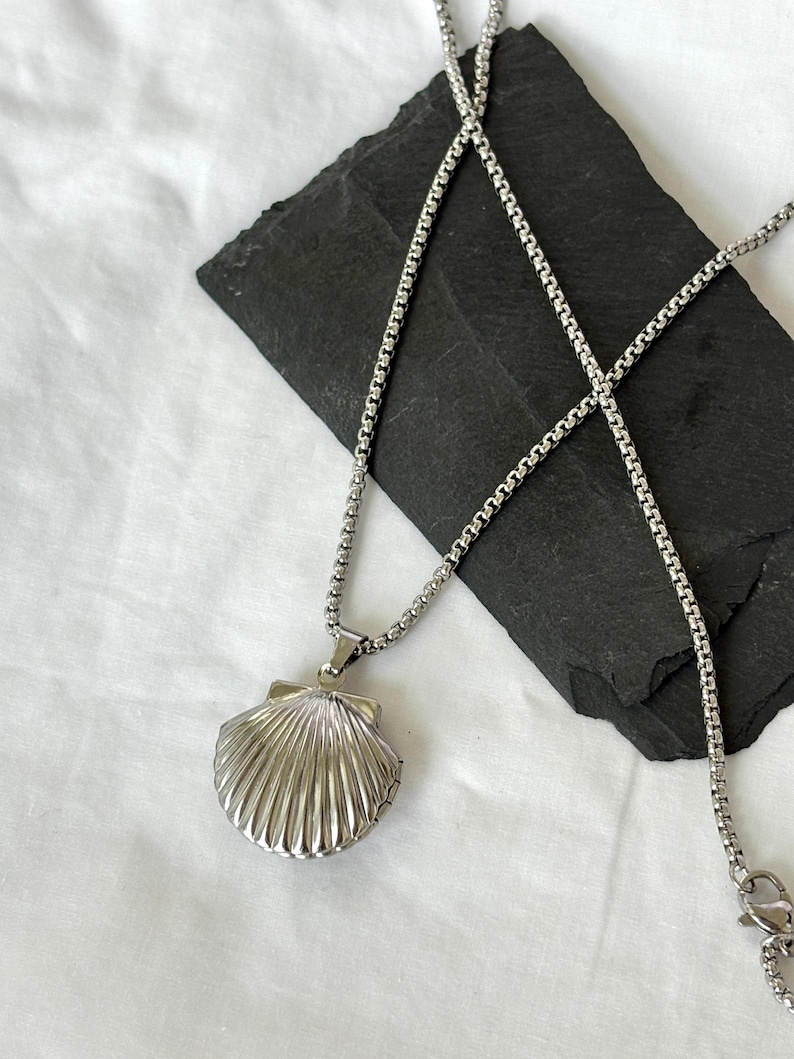 May include: A silver-colored seashell pendant necklace. The pendant is a detailed, three-dimensional scallop shell design. The necklace chain is a delicate, box-style chain. The pendant is displayed on a black, textured surface against a white background.