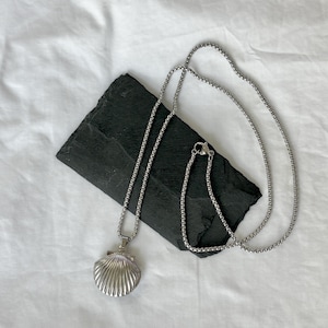 May include: A silver-colored necklace with a seashell pendant. The pendant is detailed with ridged lines, and the chain is a simple, linked design. The necklace rests on a dark, rectangular stone against a white background.