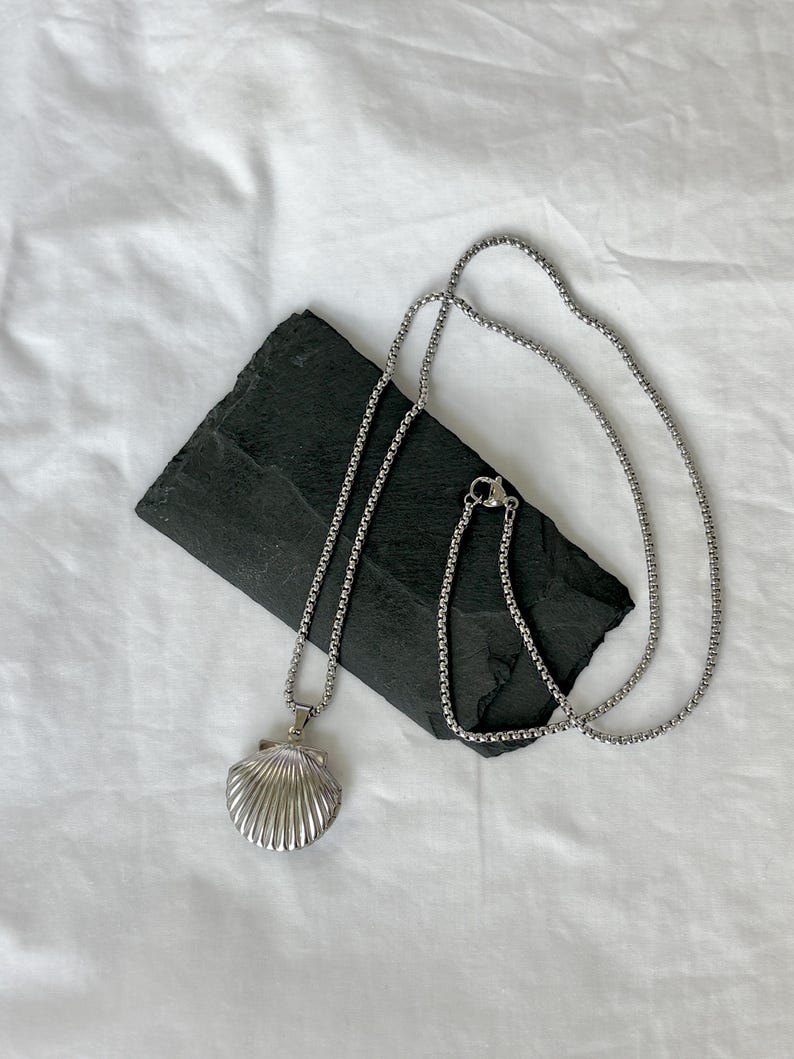May include: A silver-colored necklace with a seashell pendant. The pendant is detailed with ridged lines, and the chain is a simple, linked design. The necklace rests on a dark, rectangular stone against a white background.