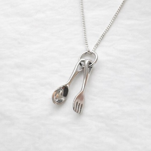 Op de afbeelding: Zilverkleurige ketting met een lepel- en vorkhanger. De hanger is bevestigd aan een fijne ketting, waardoor een minimalistisch en elegant ontwerp ontstaat. De ketting wordt gepresenteerd tegen een witte achtergrond.