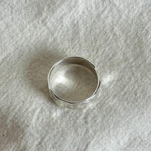 May include: A silver ring with a textured, uneven surface. The ring is circular and appears to be made of a shiny metal. The ring is displayed on a white, textured fabric.