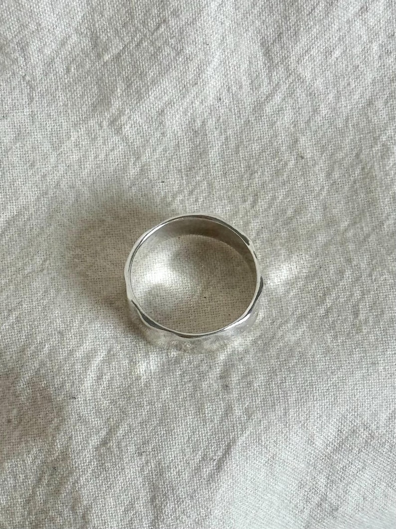 May include: A silver ring with a textured, uneven surface. The ring is circular and appears to be made of a shiny metal. The ring is displayed on a white, textured fabric.