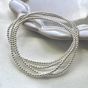 May include: Four silver beaded bracelets are arranged in a circular pattern on a white ceramic plate. The bracelets are made of small, round beads. The background is a light grey fabric.