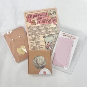 May include: A collection of jewellery and accessories, including pearl earrings, a ring in a small box, a pink polishing cloth, and a vintage card with French text. The items are arranged on a white surface.