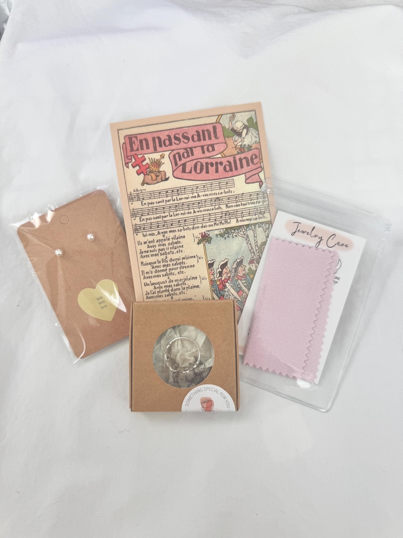 May include: A collection of jewellery and accessories, including pearl earrings, a ring in a small box, a pink polishing cloth, and a vintage card with French text. The items are arranged on a white surface.