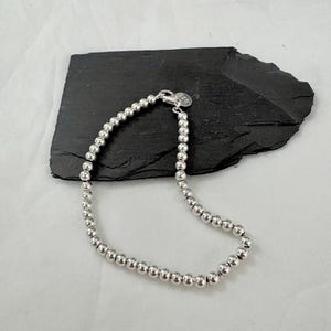 Op de afbeelding: Een zilveren kralenarmband met een karabijnsluiting. De armband bestaat uit kleine, ronde, zilveren kralen. De sluiting heeft een kleine, ronde bedel. De armband wordt gepresenteerd op een zwart, getextureerd oppervlak.