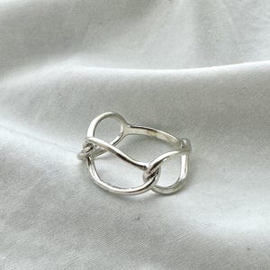 Puede incluir: Un anillo de plata con un diseño abstracto. El anillo presenta una serie de bucles ovalados interconectados, creando una estética única y moderna. El metal tiene un acabado pulido, reflejando la luz y agregando atractivo visual.