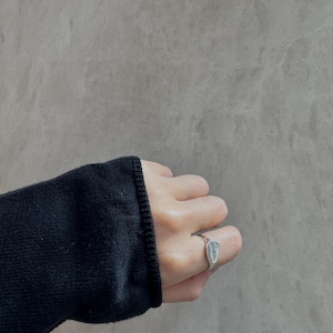 May include: A silver ring with a rectangular, textured stone is worn on a finger. The ring is set against a grey background. The person is wearing a black jumper.