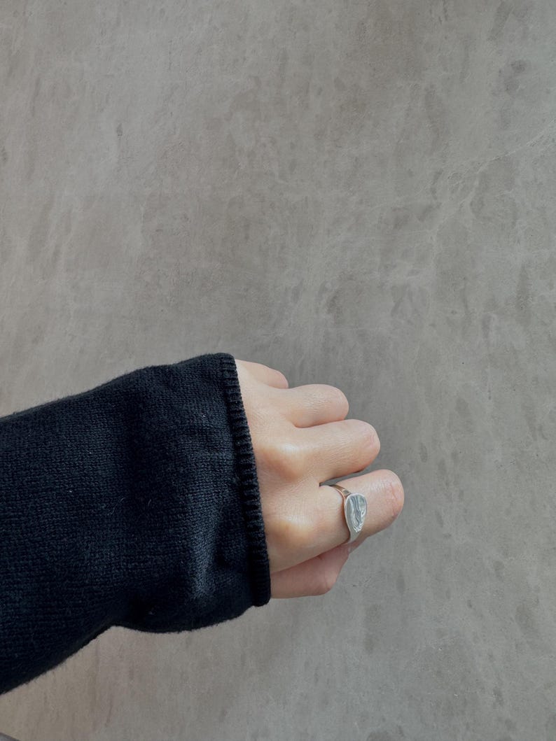 May include: A silver ring with a rectangular, textured stone is worn on a finger. The ring is set against a grey background. The person is wearing a black jumper.