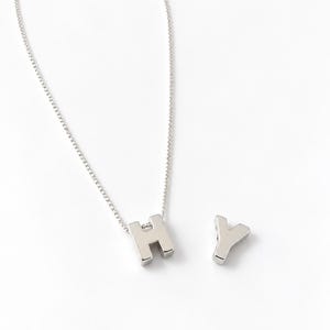 May include: A silver-toned necklace featuring a block-style letter H pendant suspended from a delicate chain, accompanied by a separate, matching silver-toned letter Y pendant placed beside it on a plain, light-coloured surface.