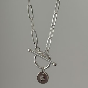 May include: A silver necklace with a toggle clasp and a small, round pendant. The pendant has the letter "B" engraved on it. The chain is made of elongated, rectangular links. The necklace is displayed against a gray background.