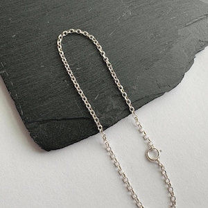 May include: A delicate silver chain bracelet with a small, circular clasp. The bracelet is composed of small, rectangular links. The jewelry is displayed on a dark gray, textured surface against a white background.
