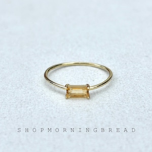 Puede incluir: Un anillo de oro con una gema rectangular de color naranja claro. El anillo está sobre una superficie de color claro. El texto "SHOP MORNING BREAD" está impreso en letras negras debajo.