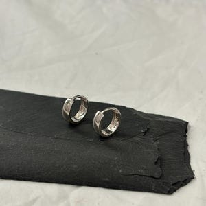 Könnte beinhalten: Zwei kleine, glänzende silberne Creolen. Die Ohrringe sind rund und haben ein schlichtes, klassisches Design. Sie werden auf einer dunklen, strukturierten Oberfläche, wahrscheinlich Schiefer, präsentiert. Der Hintergrund ist hell und neutral.