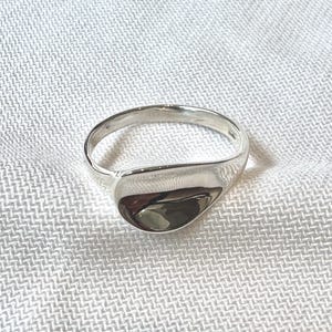 May include: A polished silver ring with a smooth, teardrop-shaped face. The ring band is a simple, rounded design. The ring is set against a white, textured background.