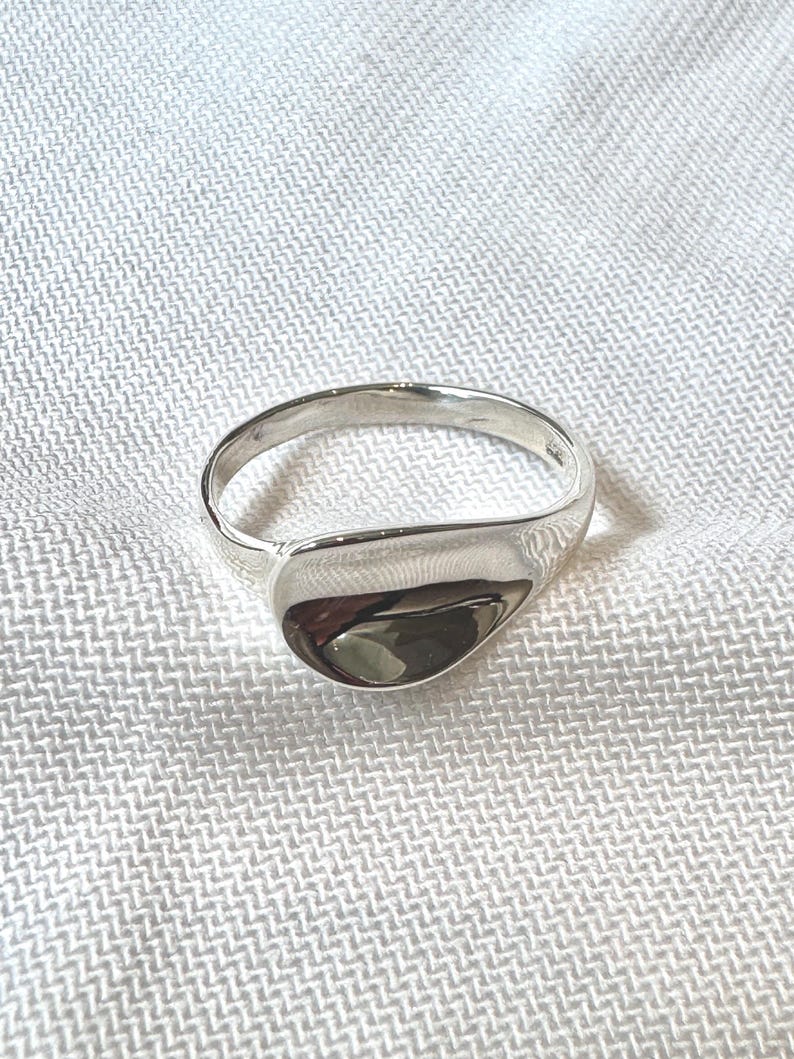May include: A polished silver ring with a smooth, teardrop-shaped face. The ring band is a simple, rounded design. The ring is set against a white, textured background.