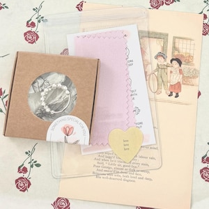 May include: A small brown box with a clear window containing a silver ring. The image includes a pink cloth, a yellow heart-shaped card with the word "love", and a vintage illustration. The box has a sticker with the text "SOMETHING SPECIAL FOR YOU".