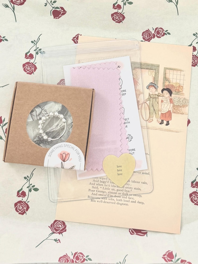 May include: A small brown box with a clear window containing a silver ring. The image includes a pink cloth, a yellow heart-shaped card with the word "love", and a vintage illustration. The box has a sticker with the text "SOMETHING SPECIAL FOR YOU".