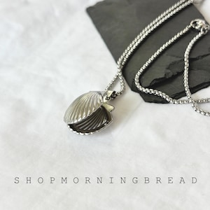 May include: A silver-colored seashell pendant necklace. The pendant is open, revealing the interior. The necklace chain is also silver. The text "SHOP MORNING BREAD" is printed at the bottom of the image.