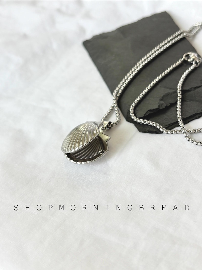 May include: A silver-colored seashell pendant necklace. The pendant is open, revealing the interior. The necklace chain is also silver. The text "SHOP MORNING BREAD" is printed at the bottom of the image.