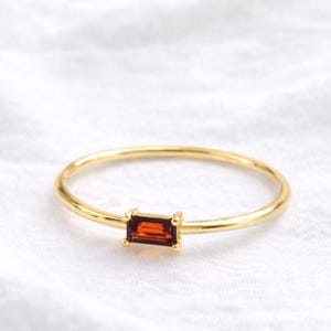 May include: A gold ring featuring a rectangular, reddish-brown gemstone. The ring's band is slender, and the gemstone is held in place by a simple four-prong setting. The ring is shown against a white, textured background.