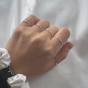 Peut inclure: Une main présentant cinq fines bagues en argent. Les bagues sont de simples anneaux, dont l'une présente un motif texturé. Un chouchou blanc avec du texte noir est visible en bas à gauche, sur un fond de tissu blanc.