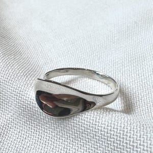 May include: A polished silver ring with a smooth, curved design. The ring has a wide, teardrop-shaped face and a simple band. The reflective surface of the ring catches the light. Jewellery.