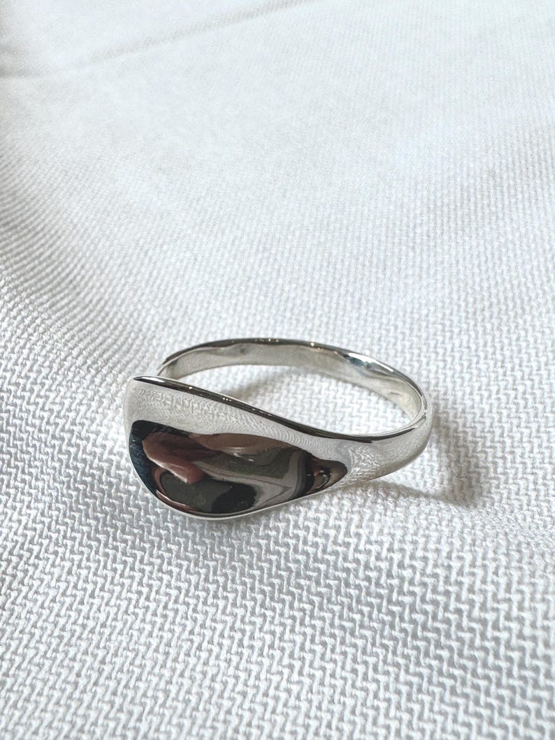 May include: A polished silver ring with a smooth, curved design. The ring has a wide, teardrop-shaped face and a simple band. The reflective surface of the ring catches the light. Jewellery.