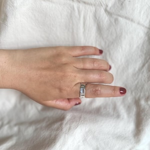 May include: A silver ring with a rectangular design is worn on a finger. The ring is made of a shiny metal and has a flat surface. The background is a white, textured fabric. The fingernails are painted a dark red colour.