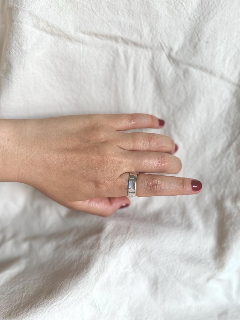 May include: A silver ring with a rectangular design is worn on a finger. The ring is made of a shiny metal and has a flat surface. The background is a white, textured fabric. The fingernails are painted a dark red colour.