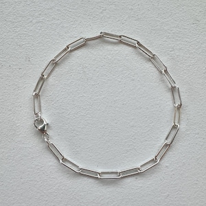 May include: A silver link chain bracelet. The bracelet is composed of a series of elongated, rectangular links. A small clasp fastens the bracelet. The jewellery is shown on a light-coloured surface.