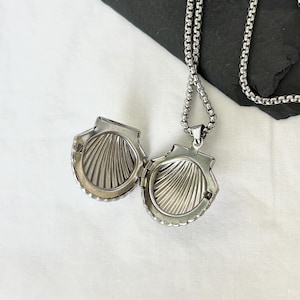 May include: Open silver seashell locket pendant on a silver chain. The locket is detailed with a ridged interior and a scalloped edge. The chain is a box chain style. The locket is open, revealing the interior.