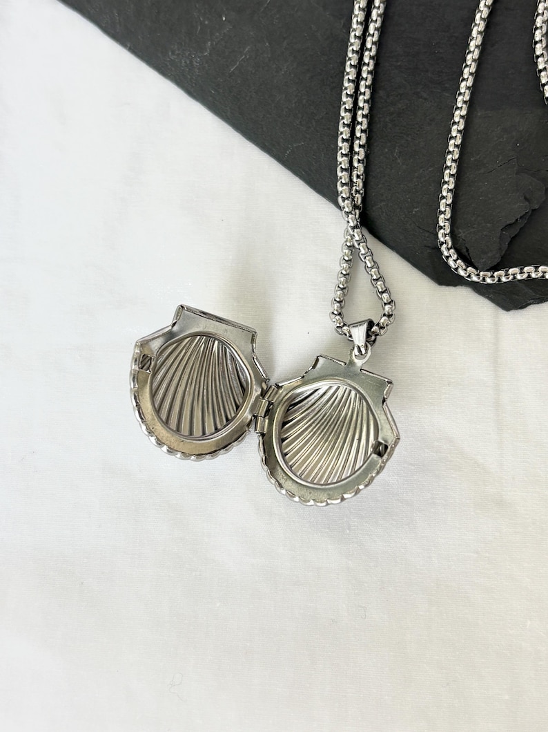 May include: Open silver seashell locket pendant on a silver chain. The locket is detailed with a ridged interior and a scalloped edge. The chain is a box chain style. The locket is open, revealing the interior.