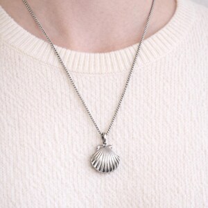 May include: A silver seashell pendant necklace. The pendant is a detailed, three-dimensional scallop shell design. The necklace chain is a delicate, linked style. The person is wearing a cream-colored sweater.